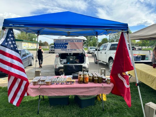 The Jarhead Treats booth: a blue canopy over a gingham table of jars and brittle, flanked by the US flag and the Marine Corps flag